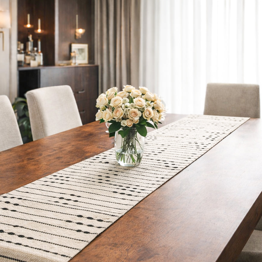 Dining table with a vase of flowers and a patterned table runner in a room with curtains and a bar area.